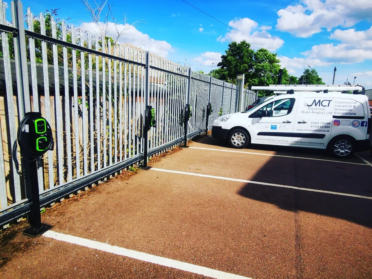Four electric vehicle charging points installed by M C T Electrical along a fenced commercial car park, with the company van alongside