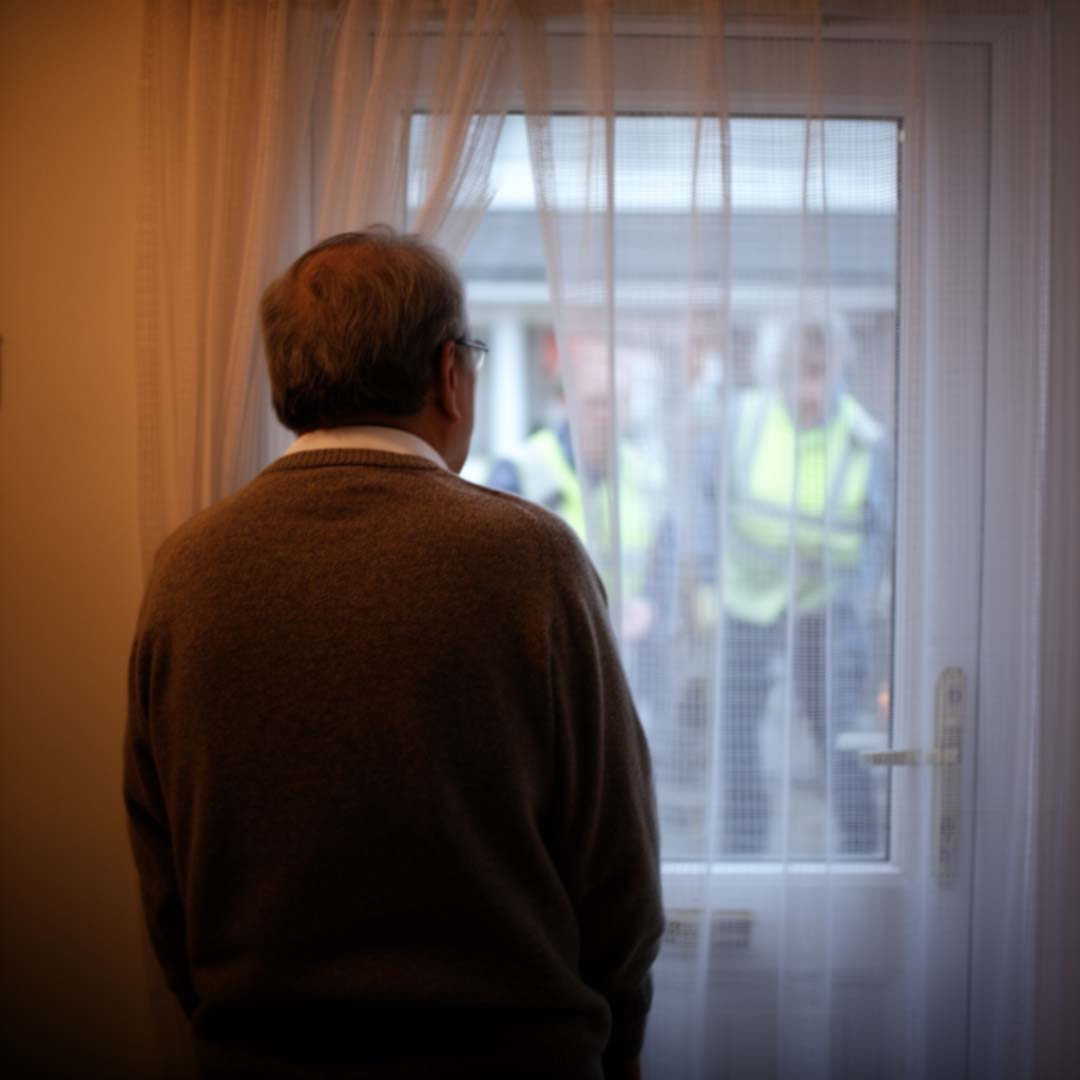 An older man cautiously looking out of his window at hi-visibility callers